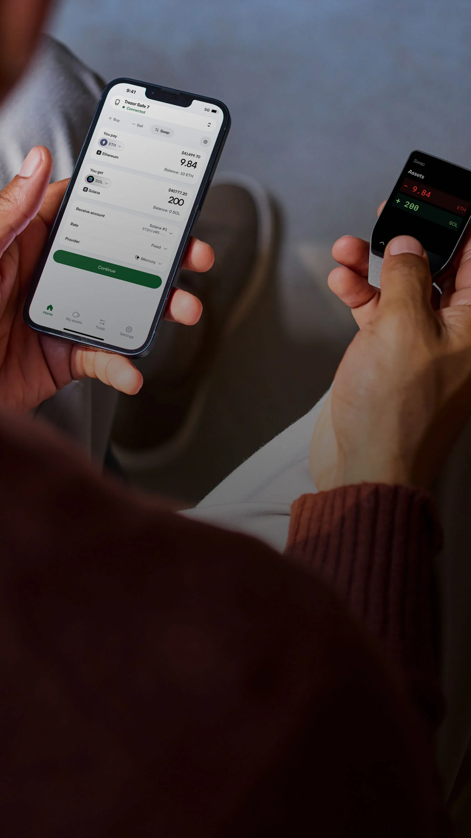 Blurry rear view of a man holding a smartphone in his left hand and a Trezor Safe 7 hardware wallet in his right hand.